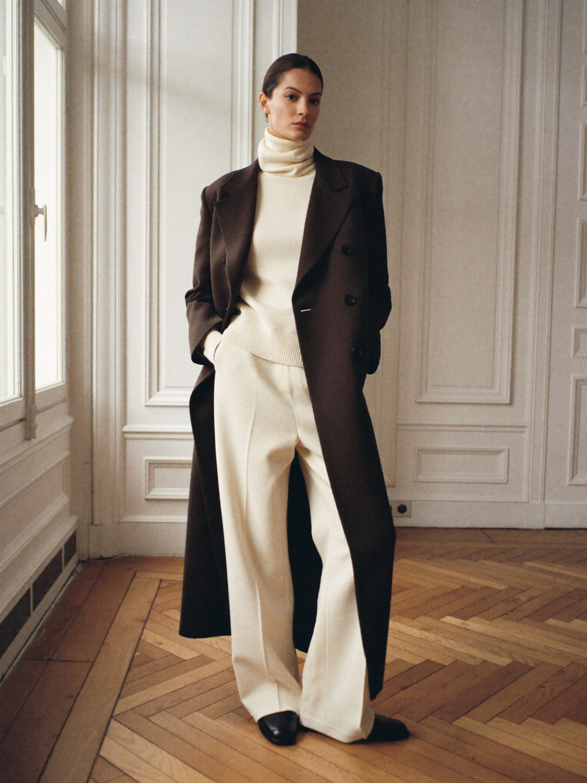 Woman in a long chocolate brown coat, cream turtleneck and cream wide-leg trousers in a paneled room